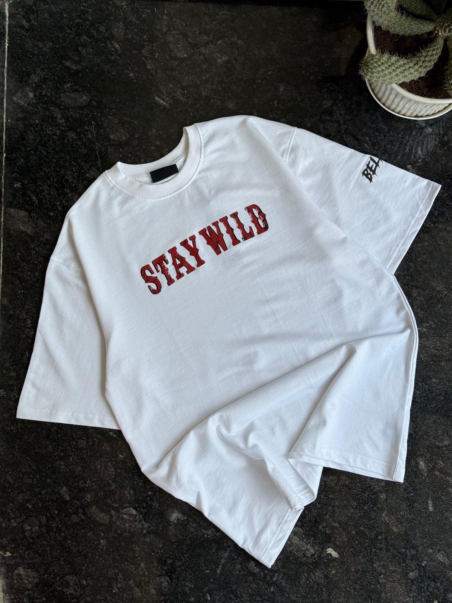 White long-sleeve shirt with 'STAY WILD' text in red on a dark surface.
